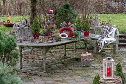 Gartentisch mit winterlicher Etagere aus Kerzen, Zapfen, Christbaumkugeln, Moos, Zuckerhut-Fichte 'Conica' (Picea glauca), Windlicht, Weidenkorb und Feuerschale im Garten