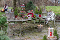 Gartentisch mit winterlicher Etagere aus Kerzen, Zapfen, Christbaumkugeln, Moos, Zuckerhut-Fichte 'Conica' (Picea glauca), Windlicht, Weidenkorb und Frau an Feuerschale