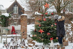 Frau vor Geschmücktem Christbaum im verschneiten Garten mit Gartenbank und Hund