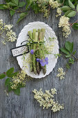 Bund grüner Spargel mit Storchenschnabel (Geranium) und Holunderblüten (Sambucus Nigra) auf weißem Teller dekoriert