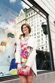 Young woman with glasses in a colorful dress and a light coat in front of a shop window