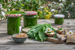 Homemade wild garlic pesto from wild garlic, pine nuts in a bowl, pesto in jars, egg, leaves on a chopping board, daisies as decoration