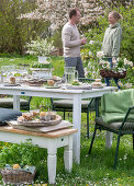 Young couple behind table laid for Easter breakfast with Easter nest and colored eggs in egg cups, daffodils and parsley in a basket in the garden