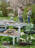 Young couple in front of a laid table, Easter breakfast with Easter nest and coloured eggs in egg cups, daffodils and parsley in a basket in the garden