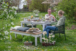 Young couple sitting at set table for Easter breakfast with Easter nest and colored eggs in egg cups, toasting with champagne, daffodils and parsley in basket in garden