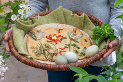 Osterfladen dekoriert mit Kräutern, gebratenen Zwiebeln und Blüte aus Paprikastreifen, Ostereier und Strauß aus Gänseblümchen im Weidenkorb mit Ostergruß