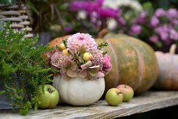 Autumnal arrangement with ornamental pumpkin vase, hydrangea flowers, dahlias, ornamental apples and apples on a wooden table