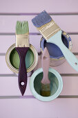 Paint brushes on cans with light green, light blue and purple paint on a wooden background