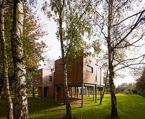 Wood-clad house on stilts with aluminium windows surrounded by trees