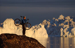 Ein Mountainbiker trägt ihr Fahrrad über Felsen, Mitternachtssonne, Jakobshavn, Ilulissat, Grönland