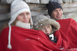 Eltern und Sohn sitzen an Holzhütte gelehnt, wärmen sich mit roter Decke