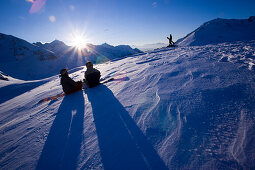 Zwei Personen sitzen mit Snowboards im Schnee, eine Person steht im Hintergrund, Kühtai, Tirol, Österreich
