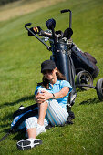 Junge Frau sitzt im Gras lehnt an Golftasche