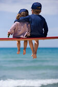 Zwei Kinder sitzen auf einer Bank am Strand, Apulien, Italien