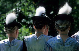 Three young women in dirndl dresses from the rear wearing traditional hats and eagle feathers, pilgrimage to Raiten, Schleching, Chiemgau, Upper Bavaria, Bavaria, Germany