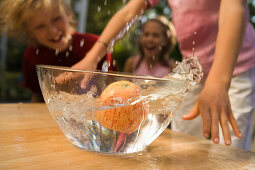 Wasser schwappt aus einer Schüssel mit einem Apfel, Kinder im Hintergrund, Kindergeburtstag