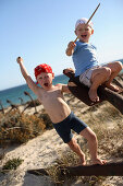 Zwei Jungen spielen Pirat auf einem rostigen Anker am Strand, Ilha de Tavira, Tavira, Algarve, Portugal