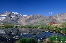 Three hikers resting at mountain lake, mountains Piz Palue and Bellavista in background, Bernina, Upper Engadin, Grisons, Switzerland