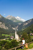 Woman resting above Heiligenblut with pilgrimage church Zum hl. Pluet, view to Grossglockner, Heiligenblut, Carinthia, Austria