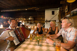 Group of tourists sitting inside a mountain hut and drinking beer while man playing accordion, Bichlalm (1731 m), Grossarl Valley, Salzburg, Austria
