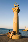 People resting on base of column with deer Elafos at port entrance of Mandraki harbour (translated literally: fold), column with doe Elafia in background, Rhodes Town, Rhodes, Greece
