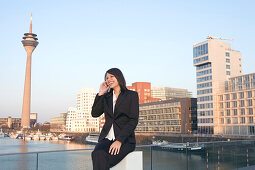 Young business woman talking on mobile phone in front of the city skyline, television tower, Zollhof, Media Harbour, architecture of Frank O.Gehry, Düsseldorf, state capital of NRW, North-Rhine-Westphalia, Germany