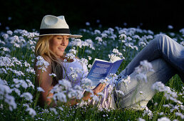 Junge Frau liegt in einer Blumenwiese und liest ein Buch, Icking, Bayern, Deutschland