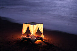 A young woman lying in a four poster bed on the beach, near Uluwatu, Bali, Indonesia