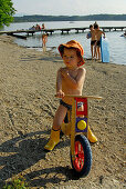 boy with sunhat on juvenile bicycle, beach of lake Hartsee, Chiemgau, Upper Bavaria, Bavaria, Germany
