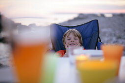 Junge sitzt am Abendtisch und lächelt, Meer im Hintergrund, Sysne, Gotland, Schweden