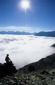 Hiker resting on rock above fogbank, Piz Julier, Upper Engadin, Grisons, Switzerland