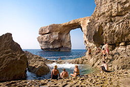 People sitting on rocks at the sea, rock arch in the background, Gozo, Malta, Europe