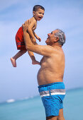 Mature man holding infant boy up at the beach