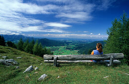 Woman sitting on bench enjoying view over valley with Inzell, alpine hut Kohleralm, Chiemgau Alps, Chiemgau, Bavaria, Germany