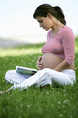 Schwangere Frau sitzt auf einer Wiese und liest