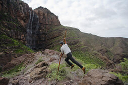 Mann beim Sprung mit kanarischem Hirtenstab vor dem Wasserfall Cascada el Escobar, Tal von El Risco, Parque Natural de Tamadaba, Gran Canaria, Kanarische Inseln, Spanien, Europa