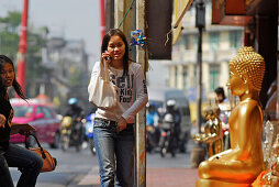 Girl with cell phone in front of Devotional Shops, Buddhas sitting and standing on the pavement, Old Town, Bamrung Muang, Bangkok, Thailand, Asia