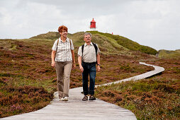 Couple walking along wooden path through dunes, lighthouse in background, Amrum island, Schleswig-Holstein, Germany