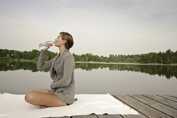 Junge Frau trinkt eine Flasche Wasser, sitzt auf einem Steg am Starnberger See, Bayern, Deutschland