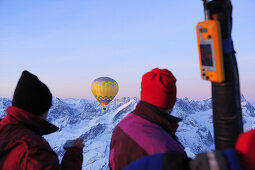 Two people on hot-air ballon ride looking towards hot-air balloon, snow covered mountains in background, Garmisch-Partenkirchen, Wetterstein range, Bavarian alps, Upper Bavaria, Bavaria, Germany, Europe