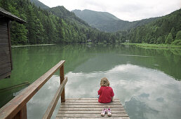 Ein kleines Mädchen sitzt auf einem Steg am See, Schaffner Weiher, Stodertal, Oberösterreich, Österreich, Alpen, Europa