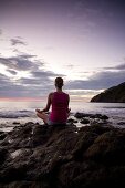 Young woman meditating on rocks at sunset in front of ocean surf at Playas del Coco, Costa Rica
