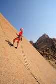 Frau seilt über rote Felsplatte ab, Große Spitzkoppe im Hintergrund, Große Spitzkoppe, Namibia