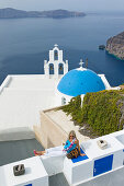Frau entspannt sich auf Mauer vor Griechisch Orthodoxer Kirche mit blauer Kuppel, Fira, Santorin, Kykladen, Griechenland, Europa