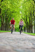 Young couple on a cycle tour, riding along a tree-lined path near Ahrensberg, Mecklenburgian Lake District, Mecklenburg-Pomerania, Germany