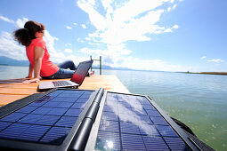 Frau sitzt auf einem Steg am Chiemsee, Laptop und Solarmodul im Vordergrund, Chiemgau, Oberbayern, Bayern, Deutschland