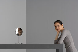 Woman sitting at table with hand under chin, smiling at camera, mirror nearby