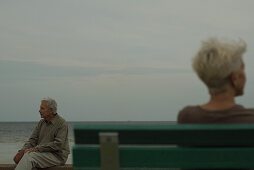 Senior man sitting on bench, looking away, senior woman in foreground