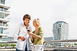 Smiling couple with mobile phone leaning against balustrade, Marco-Polo-Tower in background, HafenCity, Hamburg, Germany