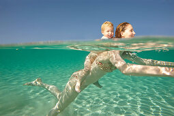 Mother giving toddler a piggyback ride. Woman swimming in crystal clear sea with young boy on her back, they are both smiling. Shot in profile from both above and below water. Underwater shot.